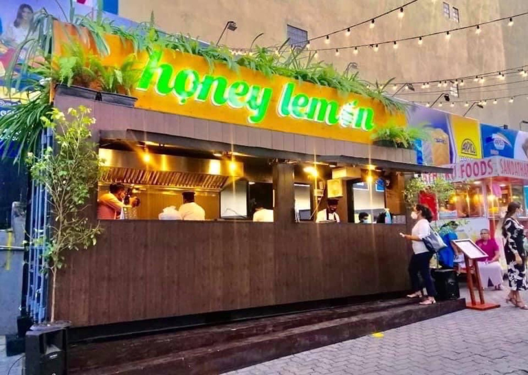 Honey Lemon container café with a green neon sign, string lights and customers