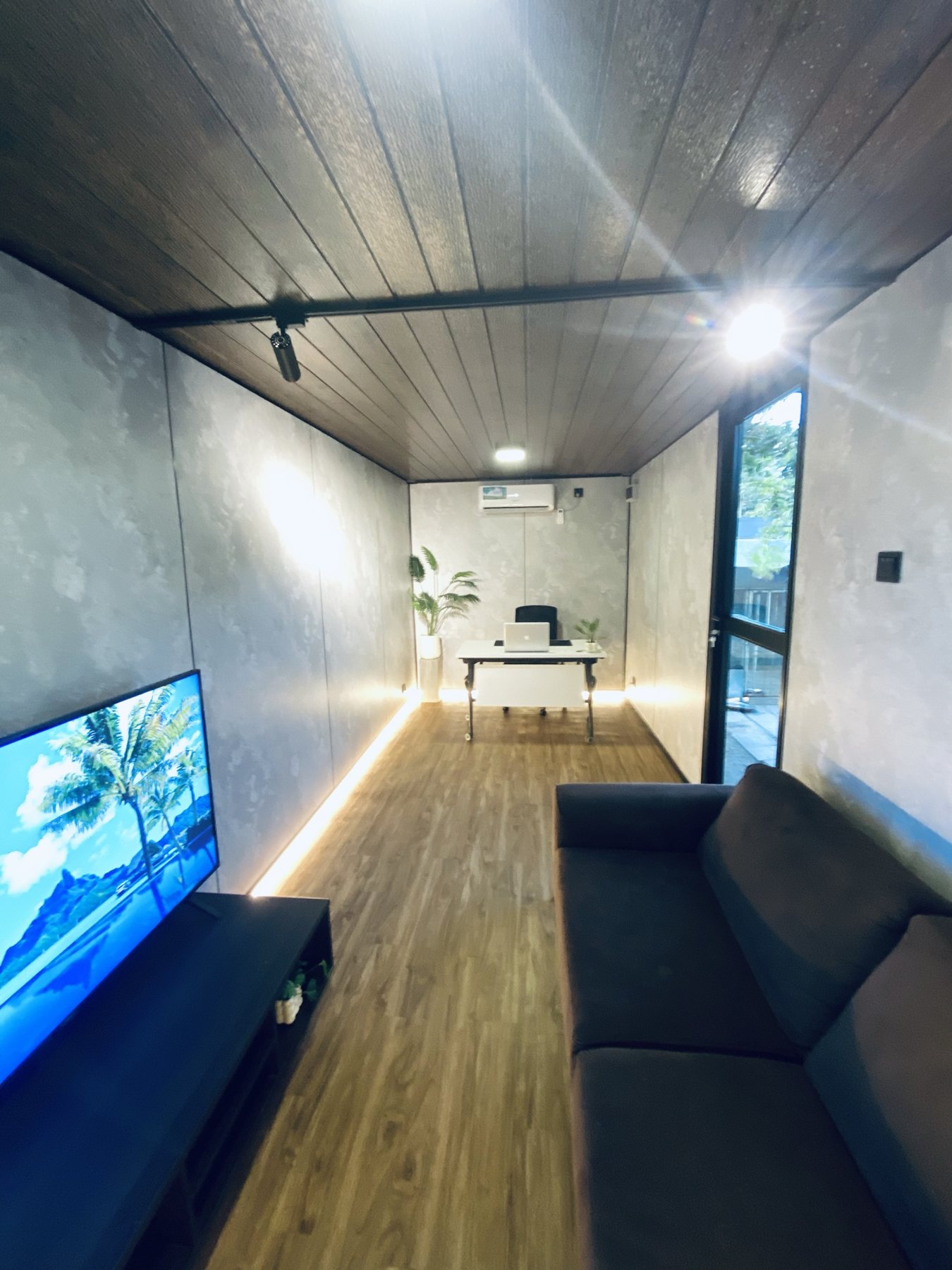 Container office interior with concrete-look walls, timber floor, sofa and wall-mounted TV